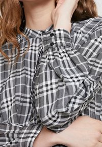 Svartvitt rutigt blus med hög krage, puffärmar och svarta knappar vid ärmans avslut, tillverkad av lätt bomullstyg.