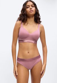 Γυναίκα με κυματιστά μαλλιά που φοράει σετ μαύρου Calvin Klein σουτιέν και εσώρουχο, στέκεται μπροστά σε απλό ανοιχτόχρωμο φόντο.