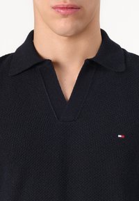 Marineblaues Sweater mit strukturiertem Strickmuster, V-Ausschnitt und Kragen. Mit einem kleinen Logo in Rot, Weiß und Blau auf der linken Seite.