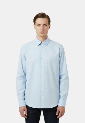 Giovane uomo che indossa una camicia azzurra con bottoni, colletto appuntito e maniche lunghe, in piedi contro uno sfondo bianco semplice.
