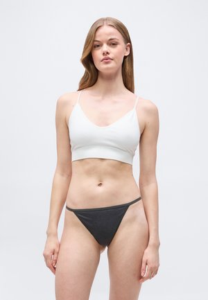 Νεαρή γυναίκα με μακριά μαλλιά, που φοράει λευκό λογκοκαμωμένο τοπ και σκούρα γκρι κάτω μέρος μπικίνι, όρθια σε απλό φωτεινό φόντο.