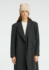 Grijs wollen dubbelzijdig jasje met geknipte revers, gecombineerd met een beige gebreide beanie. Het heeft grote knopen en lange mouwen.