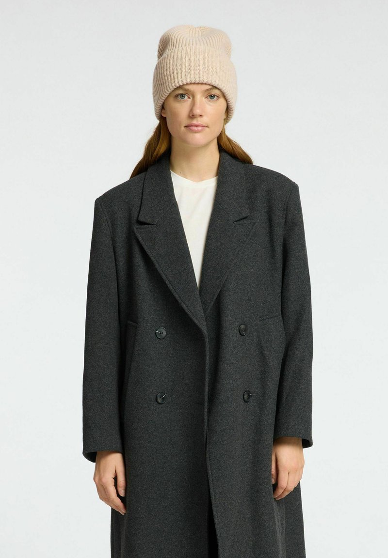 Grijs wollen dubbelzijdig jasje met geknipte revers, gecombineerd met een beige gebreide beanie. Het heeft grote knopen en lange mouwen.