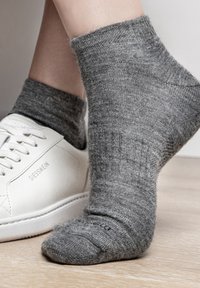 Graue Strickcrew-Socke mit gerippter Textur, getragen mit einem weißen Sneaker auf einem Holzboden. Die Socke hat eine verstärkte Zehen- und Fersenpartie.