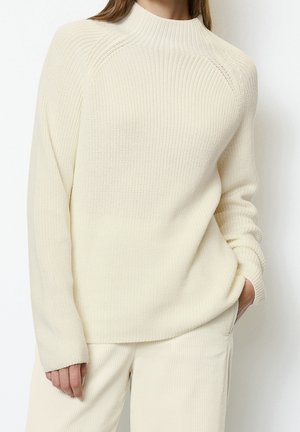 Femme portant un pull en maille côtelée crème avec manches raglan et pantalon texturé crème, debout avec une main dans la poche.