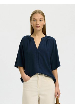 Nő, laza sötétkék V-nyakú blúzát és világos bézs nadrágját viseli, egyik kezével a zsebében áll, és egy barna pántos táskát tart.