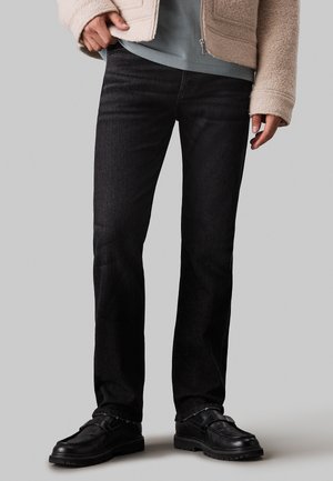 Persoon die zwarte straight-leg jeans draagt, zwarte leren loafers, een grijs overhemd en een beige jas met rits tegen een grijze achtergrond.