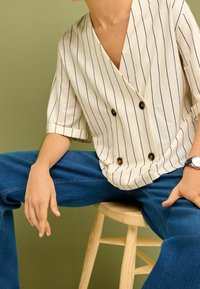 Kaksihaalarinen vaaleanbeige paita, jossa on tummansinisiä raitoja, lyhyet hihat ja käärityt hihansuut. Yhdistetty sinisiin farkkuihin.