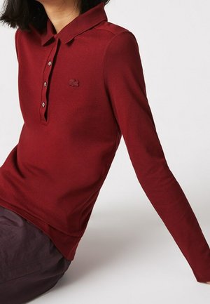 Frau trägt ein langärmeliges burgunderrotes Poloshirt und sitzt mit dem ausgestreckten rechten Arm vor hellem Hintergrund.