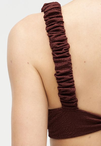 Gros plan sur une épaule nue avec une large bretelle en tissu brun foncé froncé provenant d'un vêtement texturé.