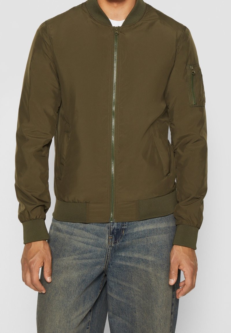 Olivově zelená bomber bunda se zipem vpředu, žebrovanými manžetami a lemem. Má boční kapsu na levém rukávu a hladký, lesklý materiál.