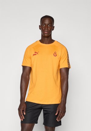 T-shirt en coton orange avec des manches courtes, un col rond et des logos rouges contrastants sur la poitrine et les manches. Associé à un short noir.