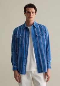 Blauwe longsleeve in een gestreept patroon. Beschikt over twee voorzakken, een knoopsluiting aan de voorkant en een kraag. Draagt over een wit t-shirt.