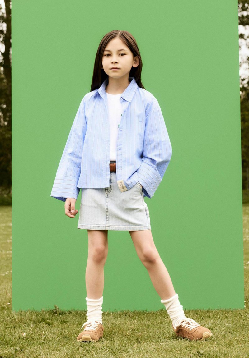 Chemise oversize rayée bleue claire, t-shirt blanc, jupe en denim, chaussettes crème, baskets beiges ; fond vert, herbe sous les pieds.