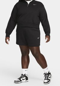 Osoba oblečená v černé mikině Nike a kraťasech, s bílými vysokými teniskami Nike a bílými ponožkami, stojí na jednobarevném pozadí.