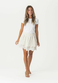 T-shirt blanc à manches courtes associé à une jupe blanche transparente à plusieurs couches, ornée de motifs floraux et d'un ourlet volanté, portée avec des talons blocs beiges.