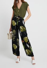 Olivgrön topp med knapprader, kombinerad med svarta vida byxor med tropisk lövtryck. Tillbehör inkluderar en vit handväska och skor.