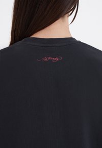 Černé bavlněné tričko s kulatým výstřihem a vyšitým nápisem "Ed Hardy" v červené barvě na zadním límci. Hladká textura.
