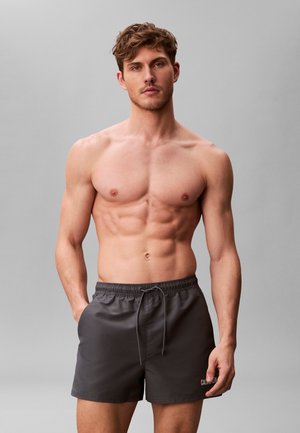 Skjorteløs mand med tonede muskler iført mørkegrå Calvin Klein badeshorts, stående med den ene hånd i lommen mod en ensfarvet grå baggrund.