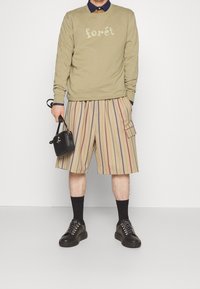 Oliwkowy sweatshirt z nadrukiem "forét", paskowane beżowe i brązowe szorty, czarna torba, czarne skarpetki i masywne czarne sneakersy.