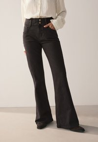 Čierne rozšírené rifle s vysokým pásom, vybavené dvomi prednými gombíkmi, bočnými vreckami a hladkou rifľovinovou textúrou. Nosia sa s krémovou košeľou na gombíky.