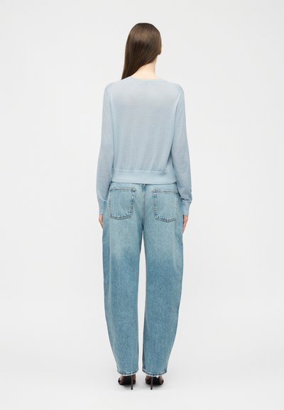 Vrouw met lang bruin haar, gekleed in een lichtblauwe trui, losse lichtblauwe spijkerbroek en zwarte hoge hakken, staand met de rug naar de kijker gericht.