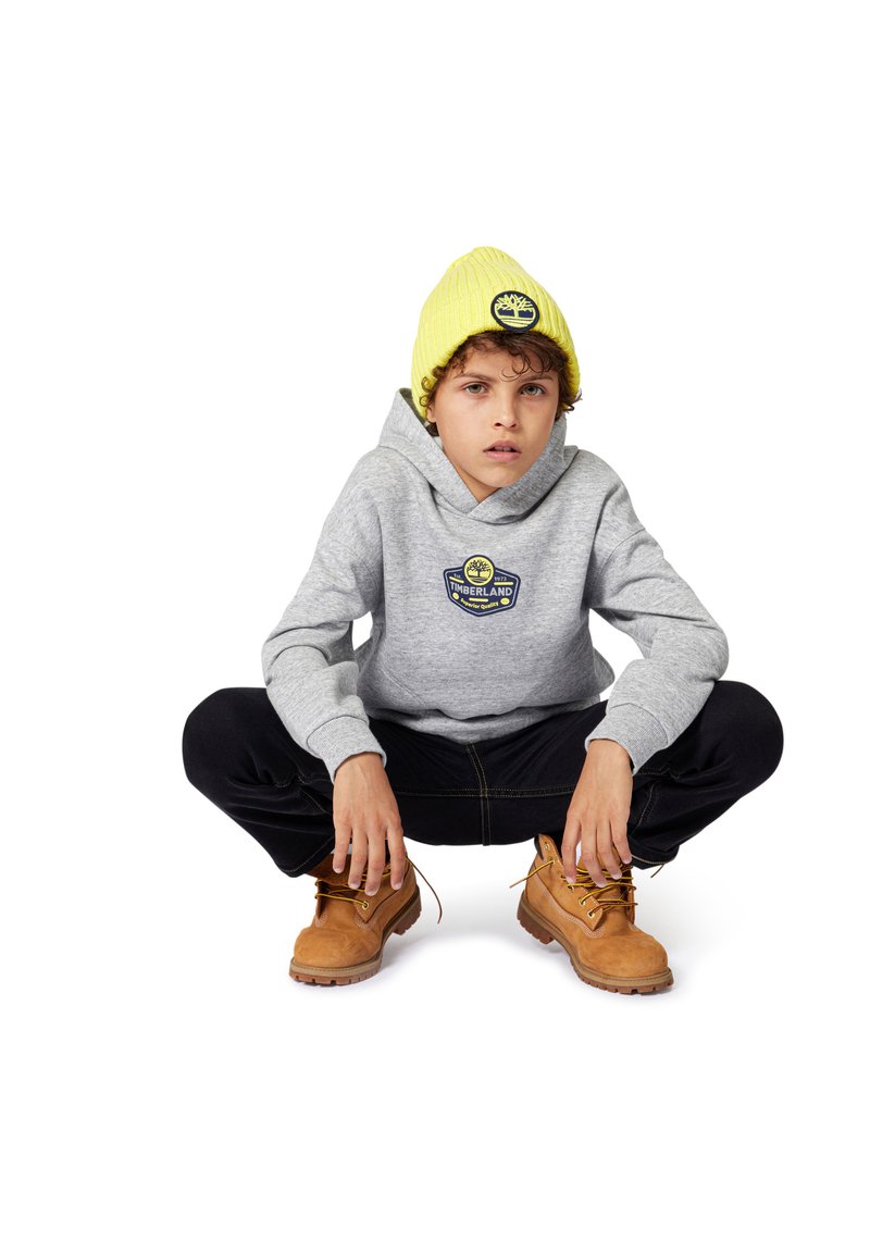 Sweat-shirt gris avec logo, pantalon noir et bottes marron. Les accessoires comprennent un bonnet tricoté jaune vif. La silhouette est accroupie avec une posture engagée.