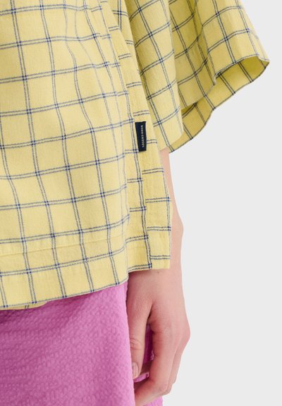 Main reposant à côté de l'ourlet d'une chemise à carreaux jaunes avec l'étiquette "Bobo Choses", au-dessus d'une jupe rose texturée sur un fond uni.