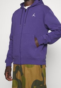 Sudadera con cremallera morada hecha de un tejido suave, con un bolsillo tipo canguro y un logo blanco en el pecho. Se ven pantalones de camuflaje por debajo.