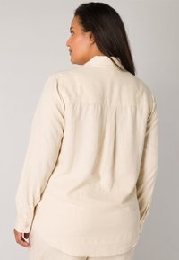 Beige long-sleeve shirt with a buttoned cuff and soft texture, featuring a yoke seam and pleat details at the back.