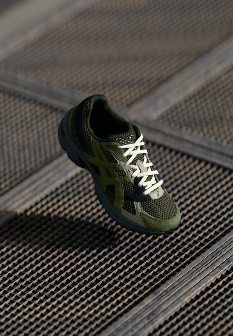 Green athletic shoe with a mesh upper, contrasting dark overlays, white laces, and a cushioned sole, positioned above a textured surface.