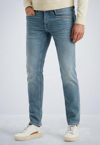 Light blue denim jeans with a slim fit, featuring five pockets and subtle fading. Paired with white sneakers and a beige sweatshirt.