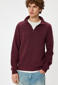 Bordeaux kleurige rits-pullover met hoge kraag, geribbelde manchetten en onderkant, gemaakt van zacht materiaal, gedragen over een wit shirt.
