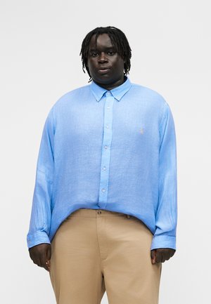 Asmuo su tamsia oda ir trumpais dreideliais, dėvintis šviesiai mėlyną marškinių tipo palaidinę ir kūno spalvos kelnes, žvelgiantis į priekį priešais paprastą foną.