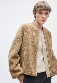 Jachetă bomber din blană sintetică bej deschis, cu captuseală netedă bej, manșete striate și fermoar frontal. Are buzunare laterale și interior matlasat.