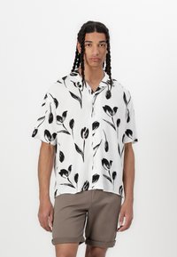 Camisa de manga corta de tela blanca con un patrón floral negro. Diseño abotonado con un corte relajado, combinada con pantalones cortos marrones doblados.