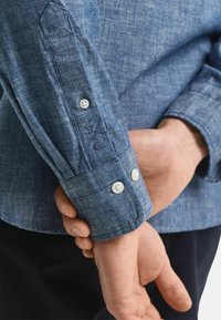 Blå chambray skjorte med lange ærmer, knapmanchetter og subtilt struktureret mønster. Hvide knapper og broderet detalje på manchetterne.