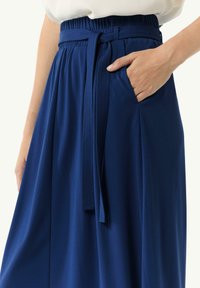 Blaues Stoffrock mit einem elastischen Bund, ausgestattet mit einem gebundenen Gürtel und einer Seitentasche. Das Material wirkt glatt und leicht.