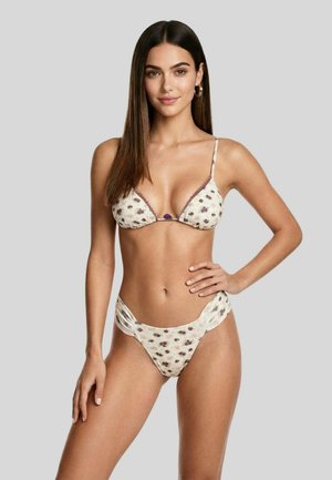 Donna con capelli lunghi e scuri che indossa un bikini floreale color crema, in piedi con una mano sui fianchi contro uno sfondo grigio chiaro uniforme.