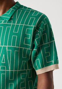 Grøn strikket polo T-shirt med et struktureret mønster af store hvide bogstaver, med kontrasterende beige kant på kraven og ærmerne.