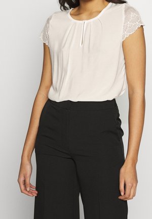 Kobieta ubrana w białą bluzkę z koronkowymi krótkimi rękawami i dekoltem z kluczem, wpuszczoną w czarne spodnie z wysokim stanem.