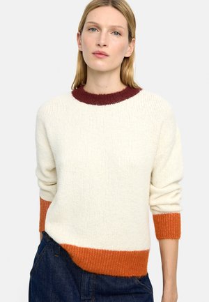 Kobieta o prostych blond włosach, ubrana w kremowy sweter z bordowym kołnierzem i pomarańczowymi mankietami, w zestawie z ciemnoniebieskimi dżinsami.