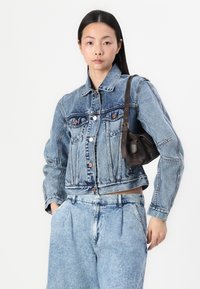 Chaqueta de mezclilla azul con un diseño corto, que presenta cierres de botón, dos bolsillos en el pecho y una superficie texturizada. Combinada con pantalones de mezclilla azul claros.