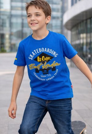 Garçon souriant portant un t-shirt bleu "Skateboarding East London", tenant un skateboard, debout sur un trottoir urbain près de bâtiments modernes.