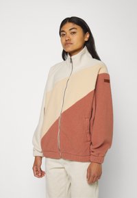Chaqueta de forro polar con cremallera en colores crema, beige y óxido, con un diseño geométrico, cuello alto y puños elásticos, que incluye bolsillos laterales.