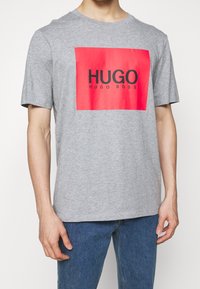 Camiseta de algodón gris con un gráfico rectangular rojo que tiene "HUGO" en texto negro y "HUGO BOSS" debajo. Cuello redondo clásico, mangas cortas.