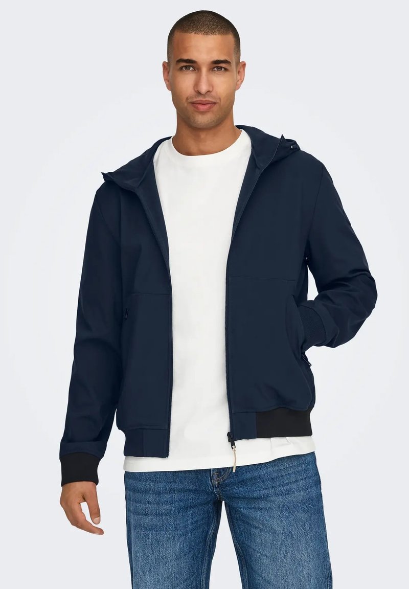 Giacca a zip blu navy con cappuccio, due tasche frontali, polsini a coste e orlo a coste, indossata sopra una t-shirt bianca e abbinata a jeans blu.