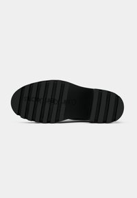 Semelle de chaussure noire avec de profondes rainures horizontales et "Calvin Klein Jeans" embossé au centre, présentée sur un fond clair uni.