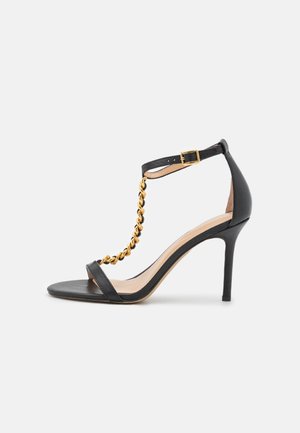 Czarne skórzane sandały na wysokim obcasie z akcentem w postaci złotego łańcucha, regulowanym paskiem wokół kostki oraz minimalistycznym wzornictwem o gładkiej teksturze.