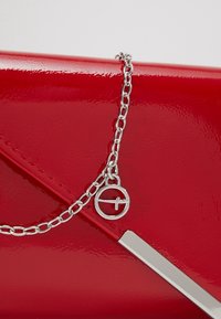 Pochette en cuir verni rouge avec chaîne argentée et pendentif circulaire, présentant un design élégant et angulaire ainsi qu'une texture réfléchissante.